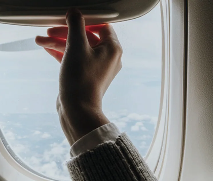 Mão ajustando a persiana da janela de um avião, com céu e nuvens ao fundo.