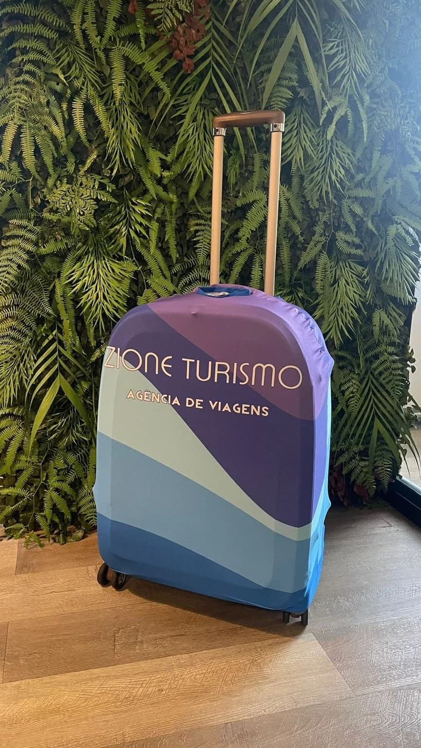 Mala com capa personalizada Zione Turismo, em tons de violeta e azul, diante de uma parede de folhagens.